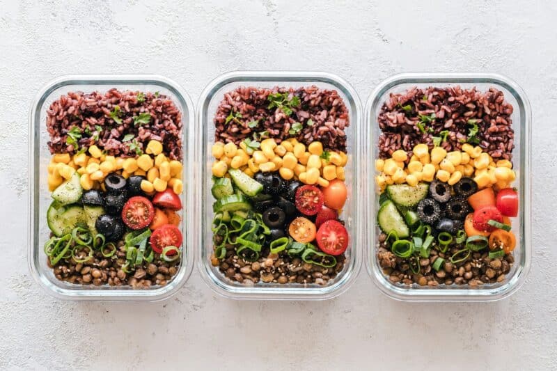 Three glass containers of healthy meals with red rice, corn, vegetables and lentils